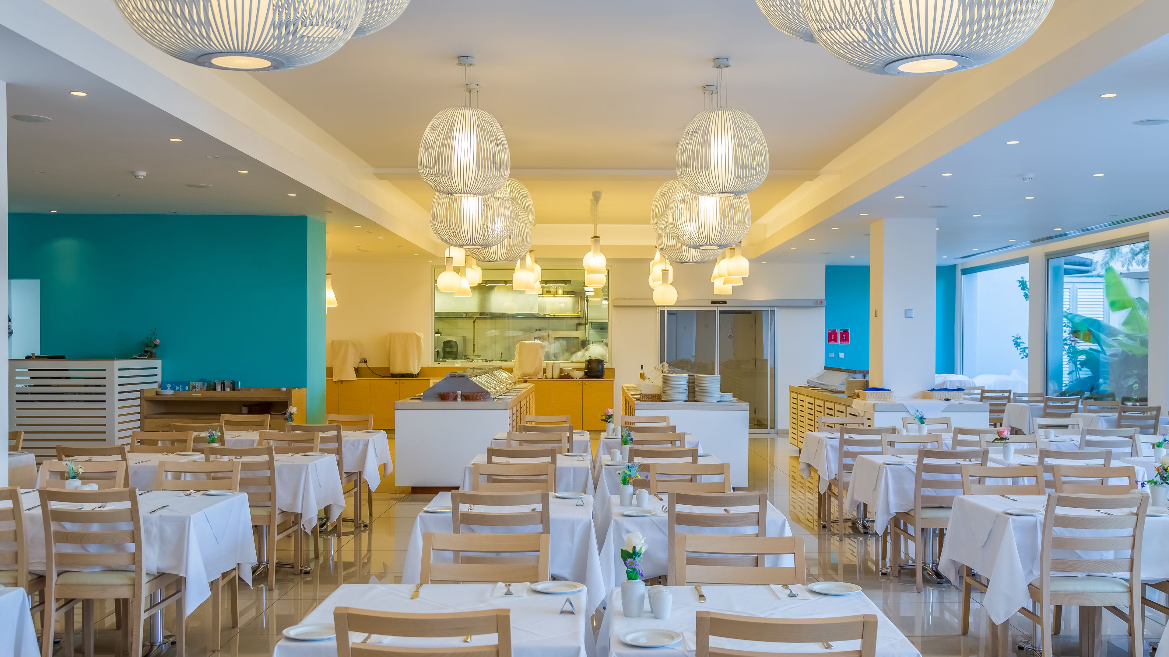 A modern restaurant with multiple dining areas, white tablecloths, wooden chairs, and hanging pendant lights.
