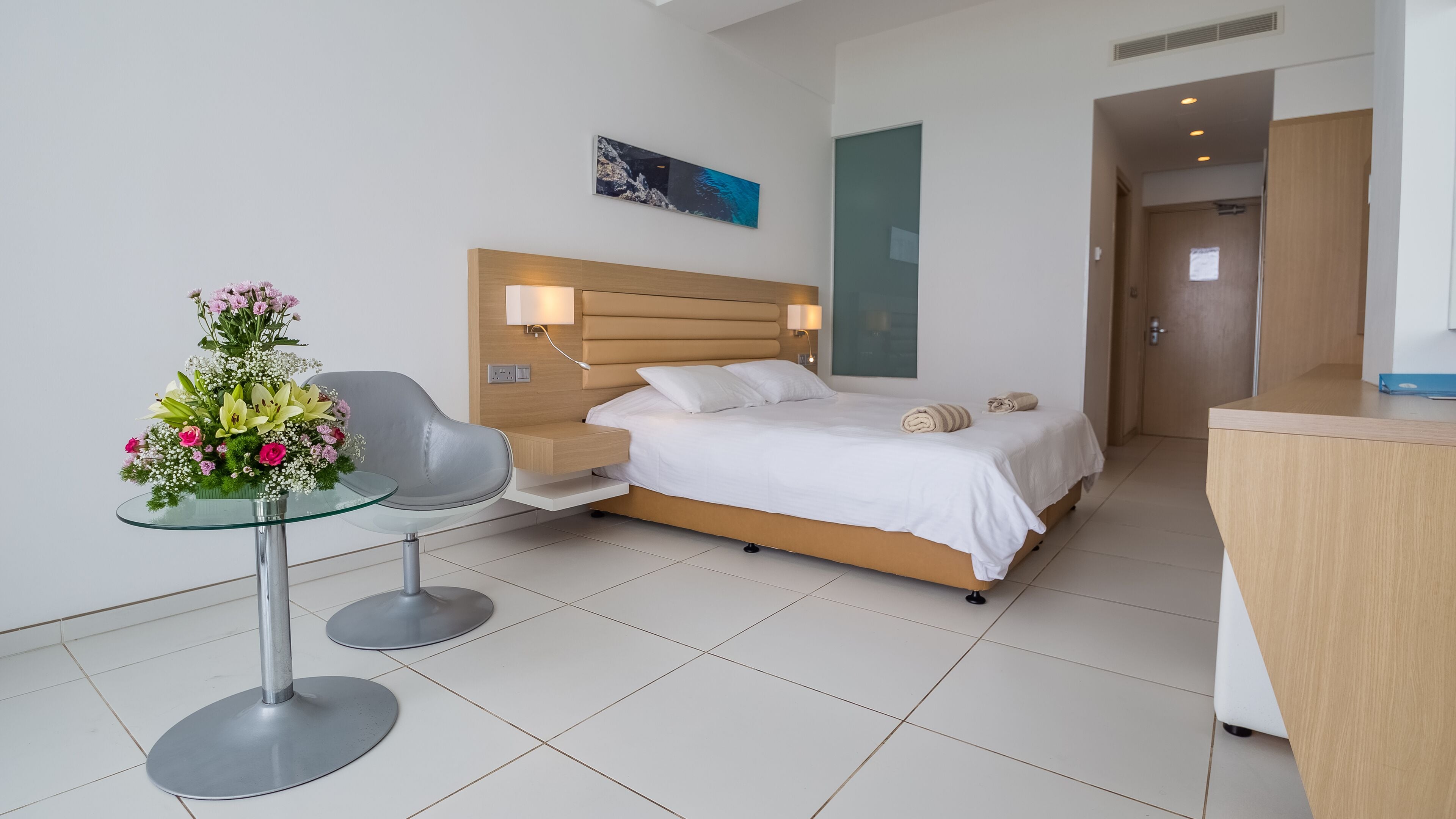 A modern hotel room with a bed, bedside tables, a desk, a chair, and a glass table with a floral arrangement.