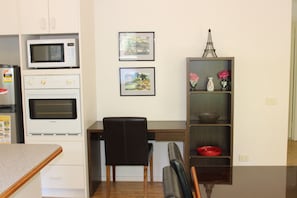 A kitchen with a microwave, oven, and refrigerator. There is a chair and a desk with a shelf containing decorative items.