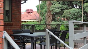 Une terrasse extérieure avec un mur en briques, un espace salon couvert avec des tables et des chaises, et une vue sur un bâtiment aux toits rouges et des arbres en arrière-plan.
