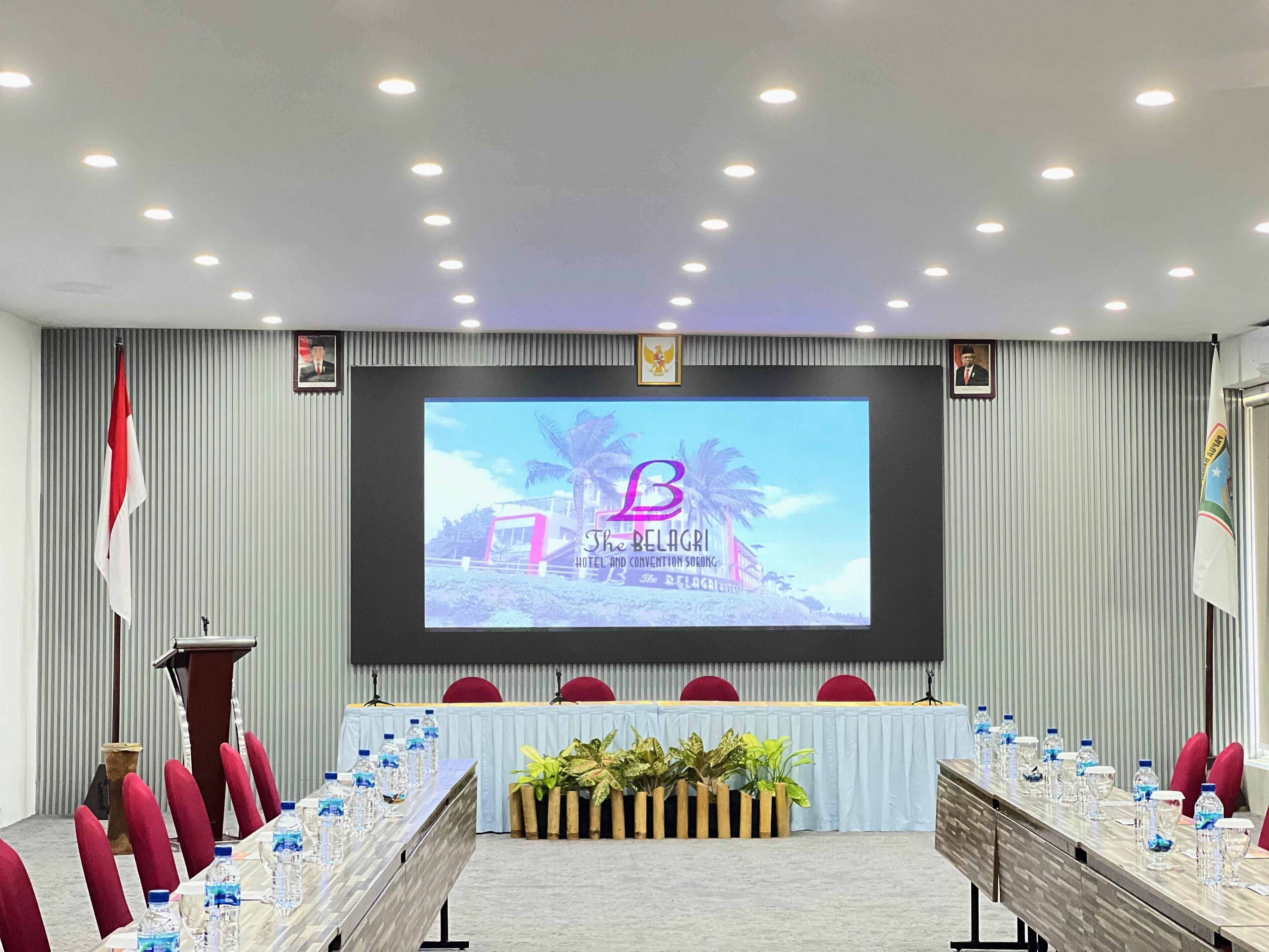 A conference room with a long table, red chairs, and a screen displaying 'The Belagri Hotel and Convention Center'.