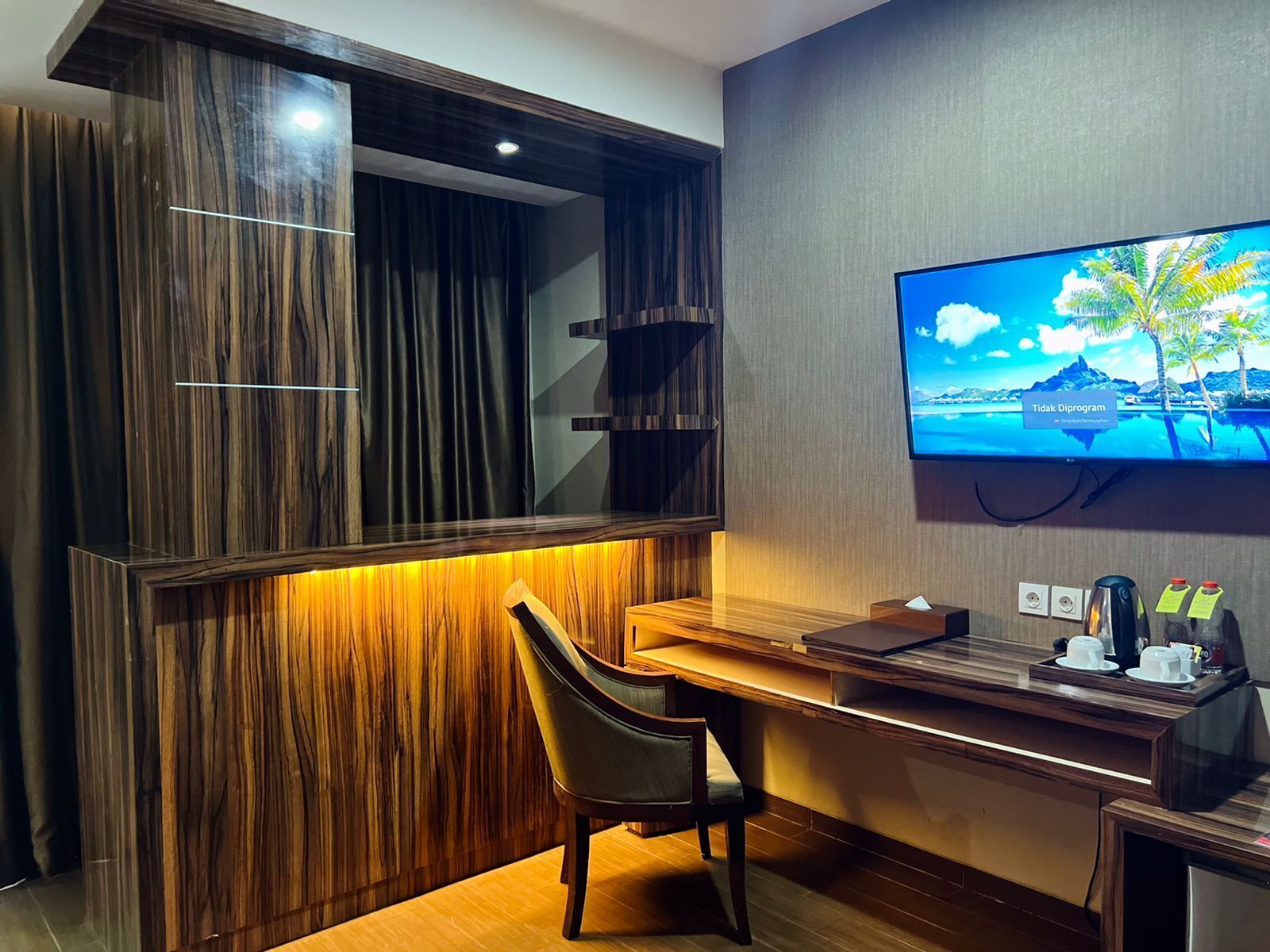 Et moderne hotelværelse med skrivebord i træ, en behagelig stol, et fladskærms-tv på væggen og et minikøleskab med kaffemaskine og glas.
