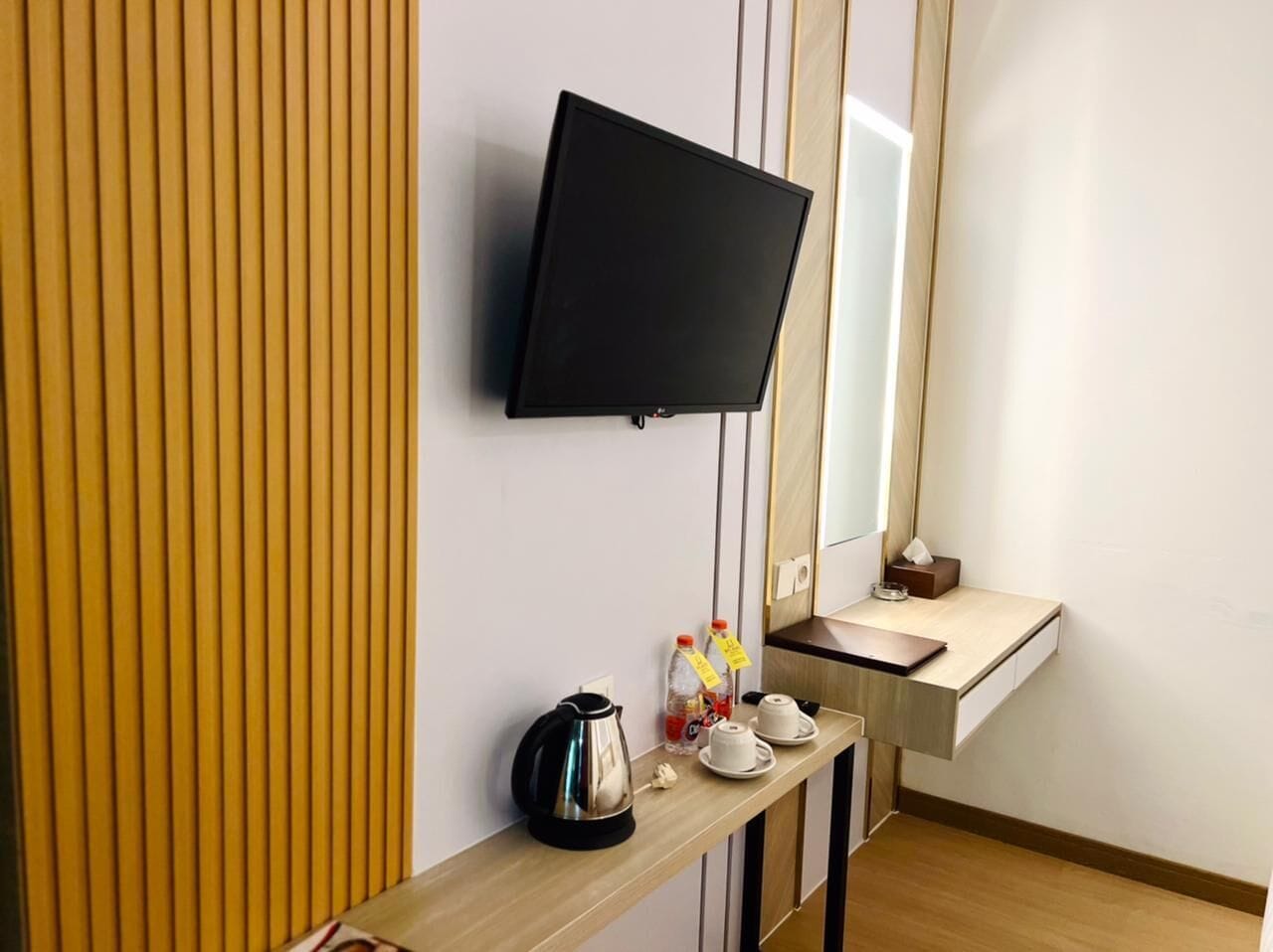 Una habitación de hotel con un televisor montado en la pared, un panel de pared de madera, una pequeña mesa de madera con una tetera, tazas y una botella.