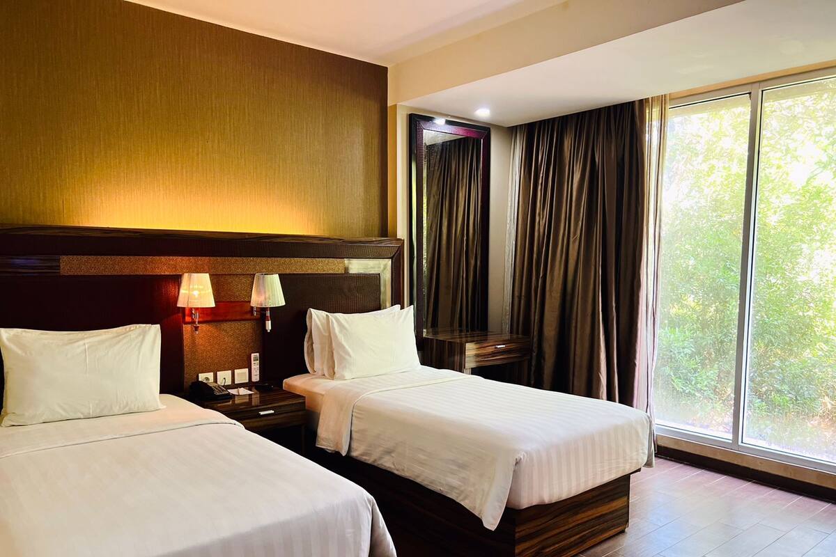 A hotel room with two beds, wooden headboards, a nightstand with a lamp, and a large window with curtains.
