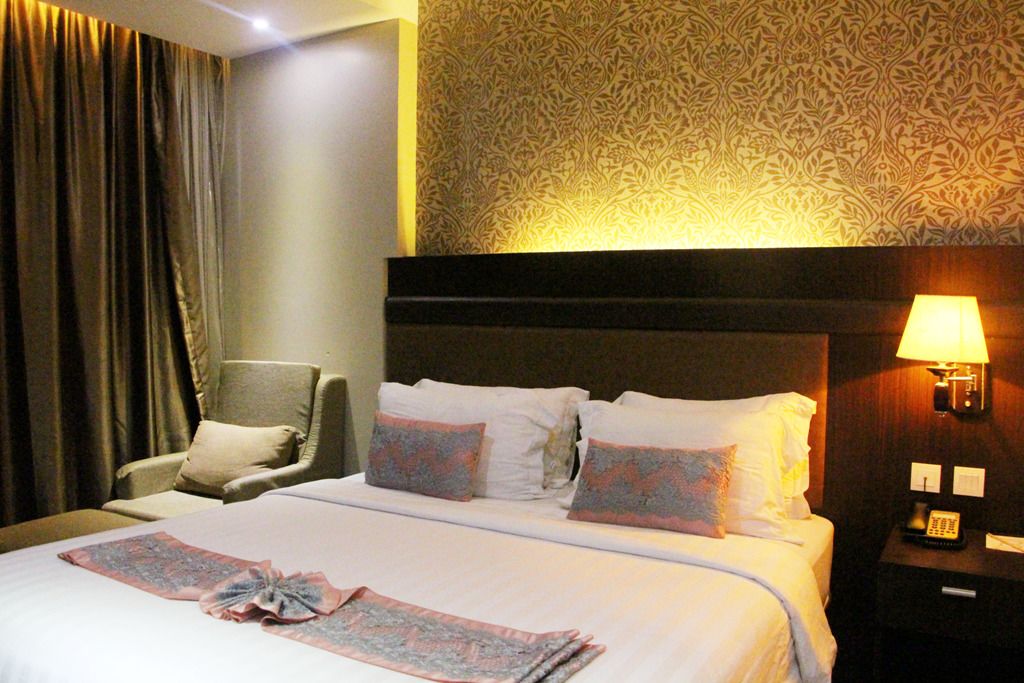 A hotel room with a bed, a chair, a nightstand with a lamp, and a patterned wallpaper.