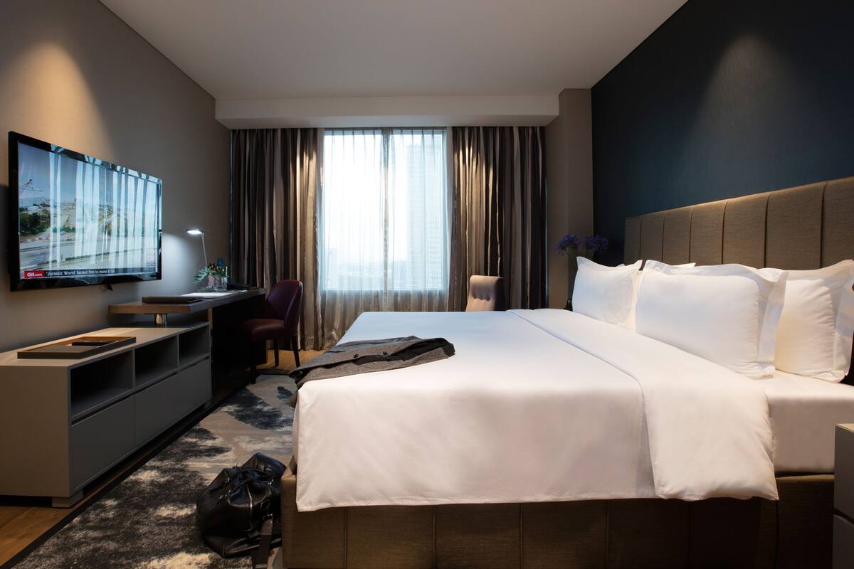 A modern hotel room with a large bed, a desk, a TV, and a view of the city through the window.