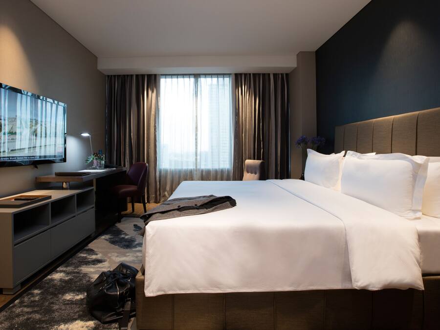 Habitación de hotel moderna con una cama grande, un escritorio, un televisor y vistas a la ciudad desde la ventana.