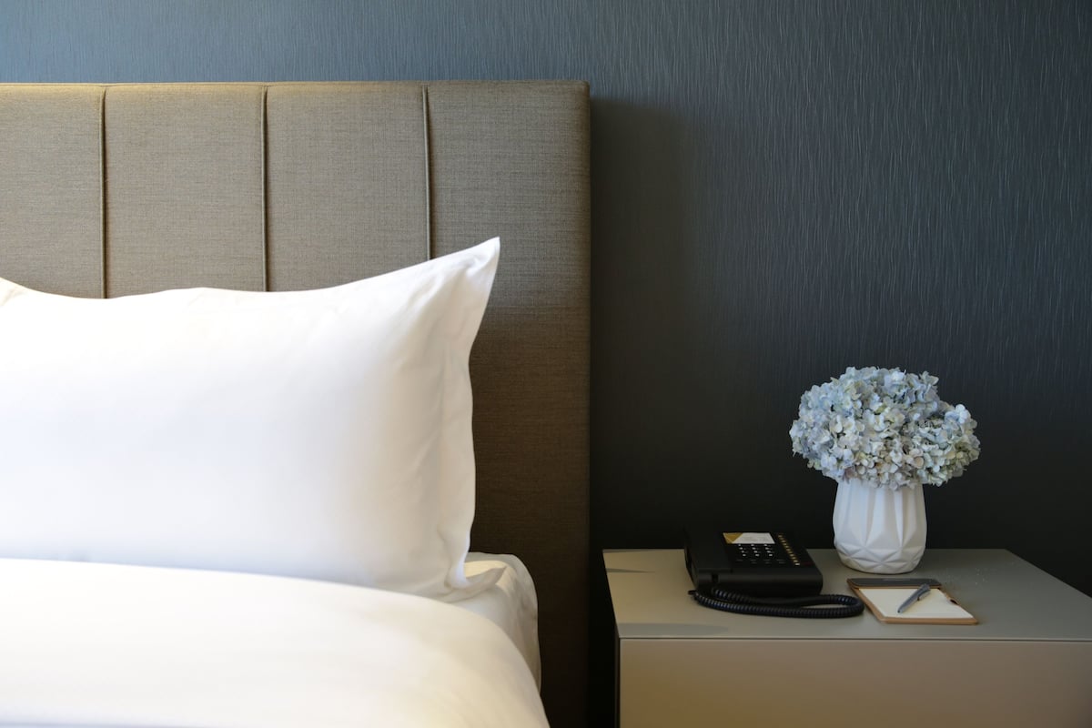 A neatly made bed with white bedding, a bedside table with a phone, a vase with flowers, and a small notepad.