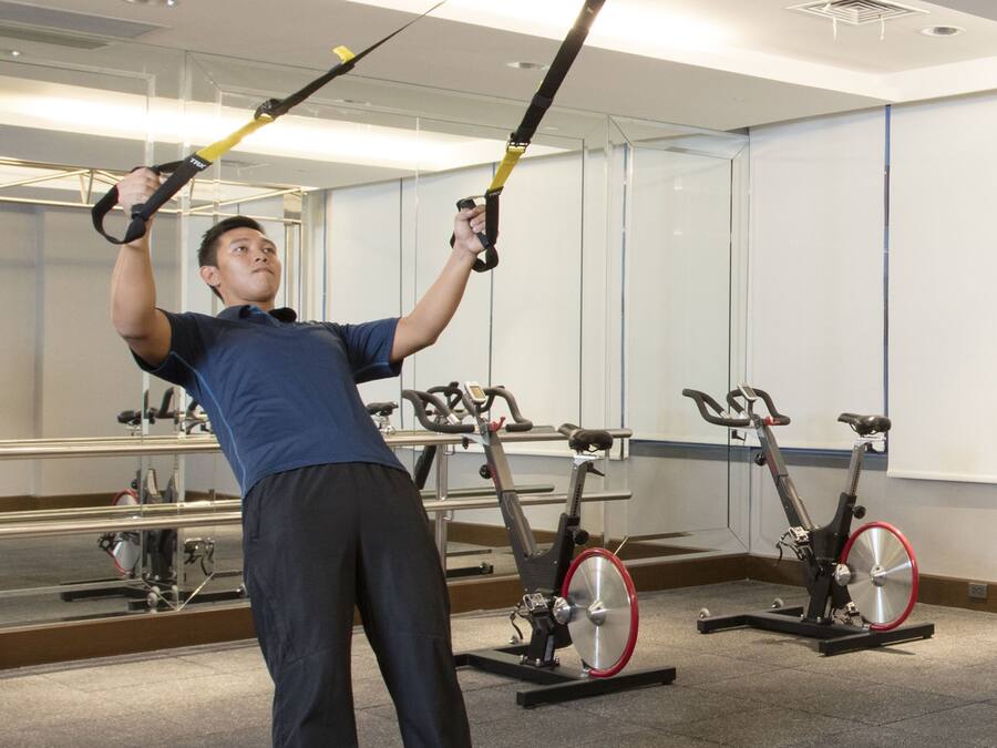 A man exercising with TRX handles in a gym with stationary bikes.