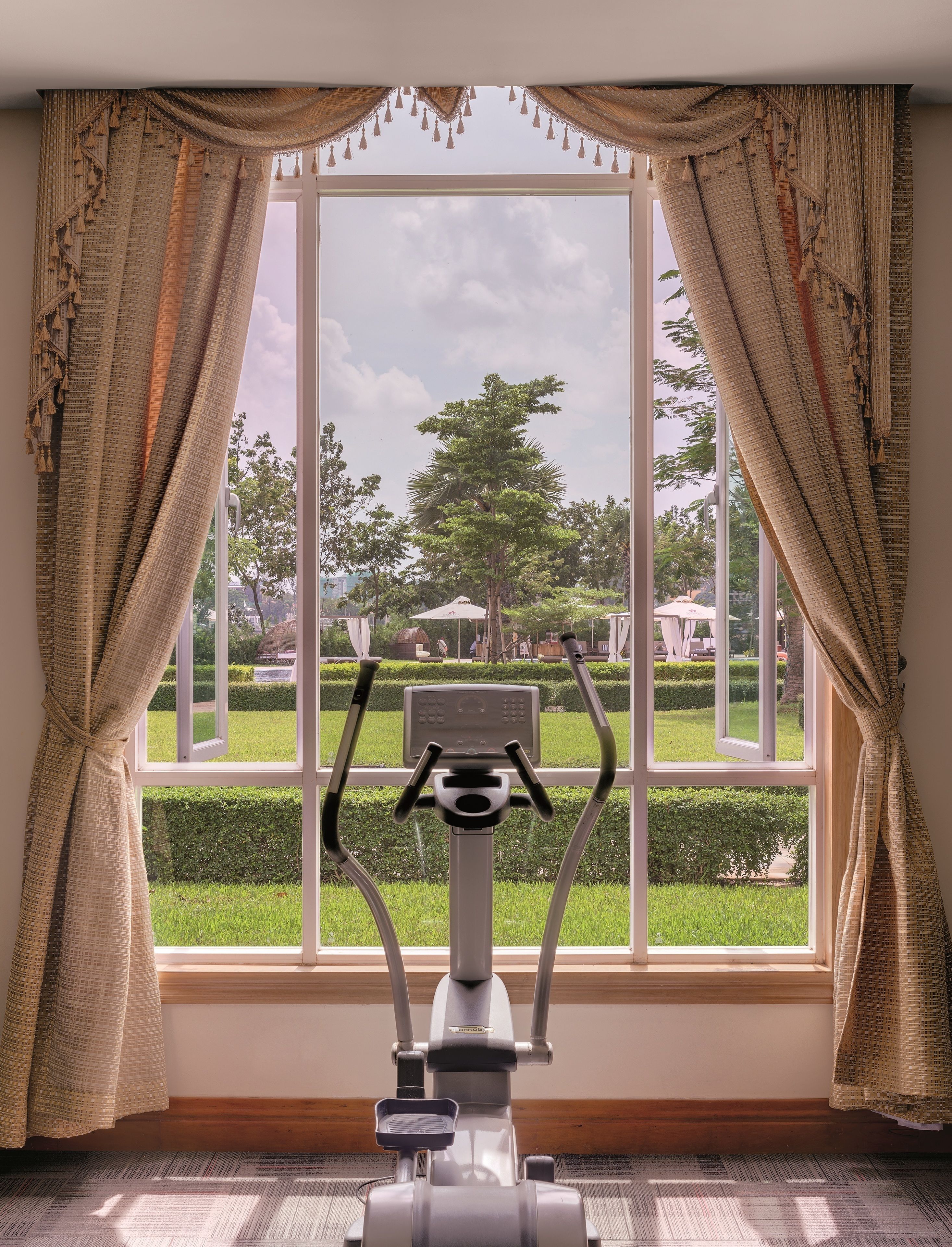 An exercise bike in a room with large windows, curtains, and a view of a park.
