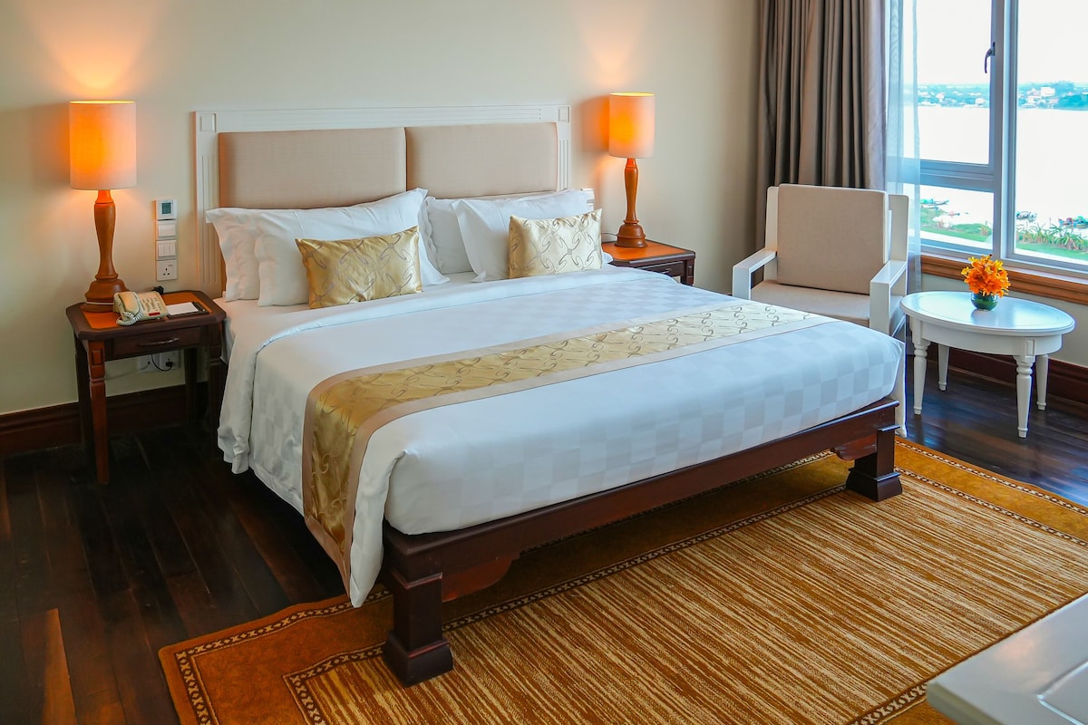 A hotel room with a large bed, bedside lamps, a small table with a vase, and a view through the window.