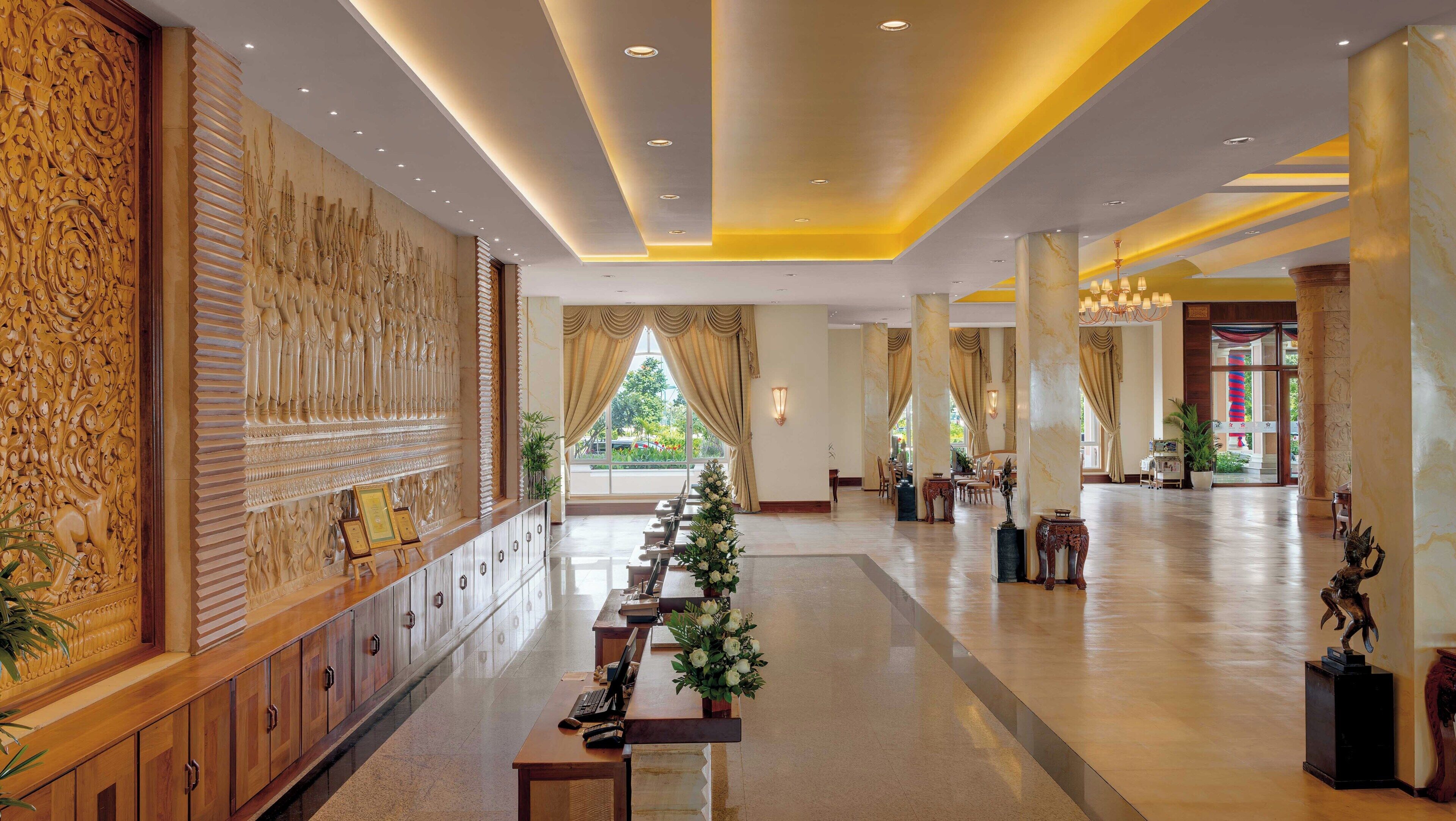 A spacious lobby with a long reception desk, a decorative wall with intricate carvings, and a large window with curtains.