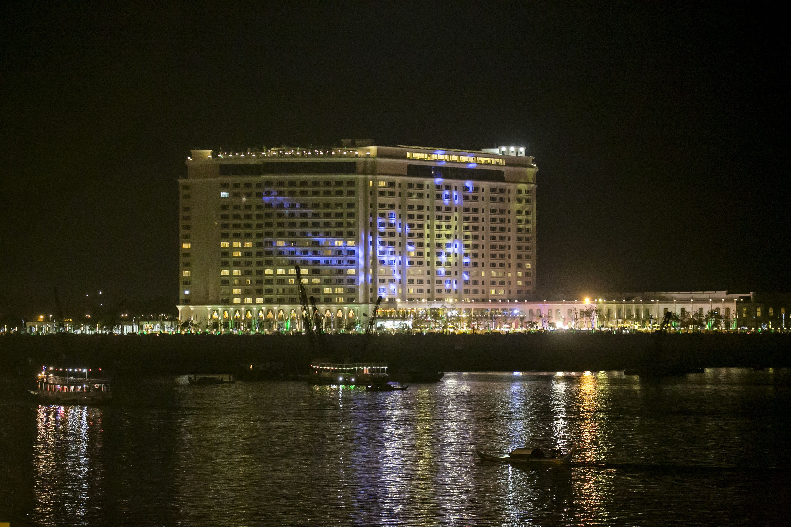 A large hotel with multiple floors and balconies, situated by a waterfront with boats and a bridge in the background.