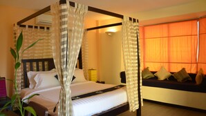 A four-poster bed with a canopy, a bedside table, a nightstand, a window with curtains, and a bench with cushions. - Robam Inn (Siem Reap)