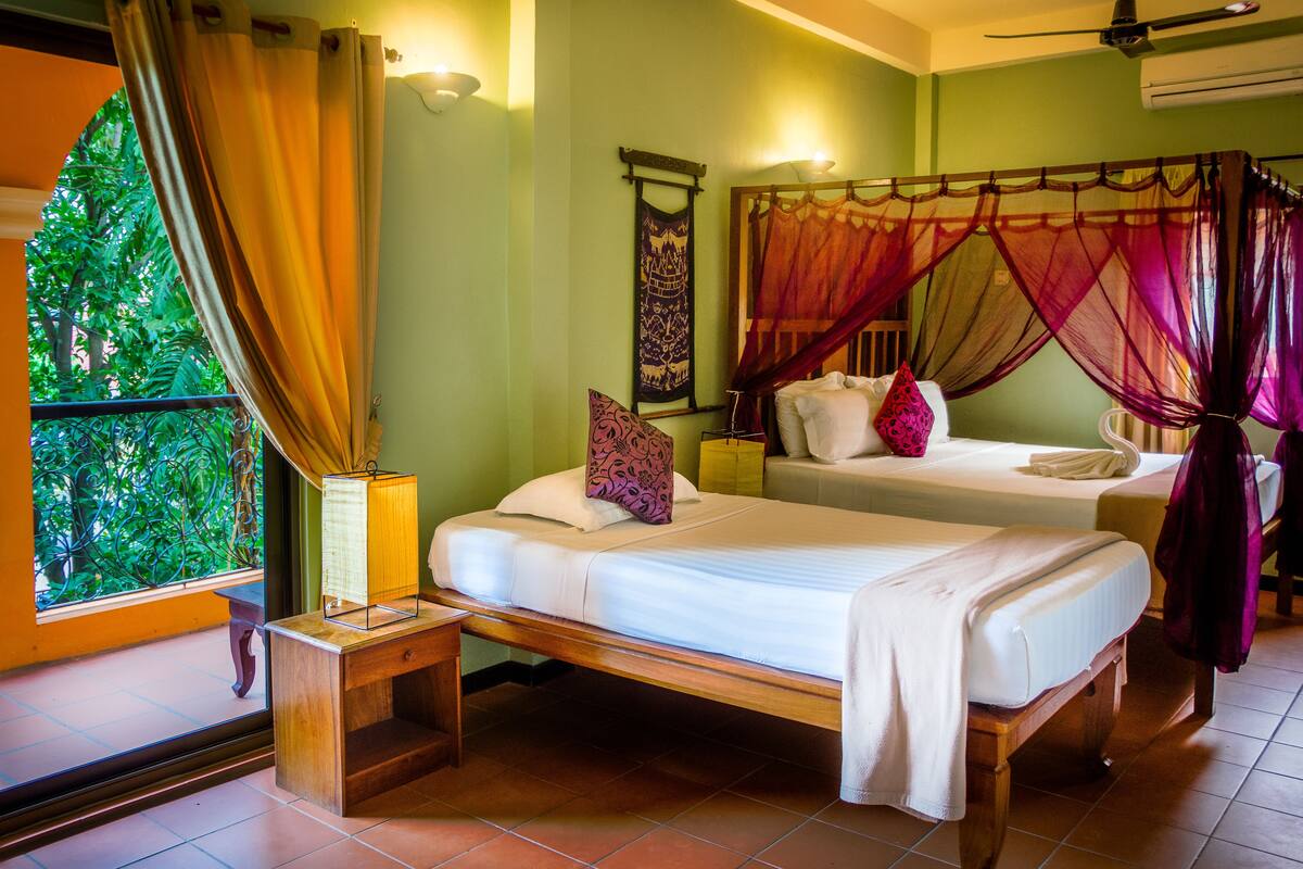 A room with two beds, a window with curtains, a bedside table, and a wall-mounted air conditioner.
