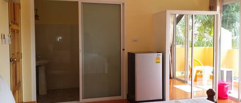A bedroom with a bed, a sliding glass door leading to a balcony, a small refrigerator, and a bathroom with a toilet and sink.