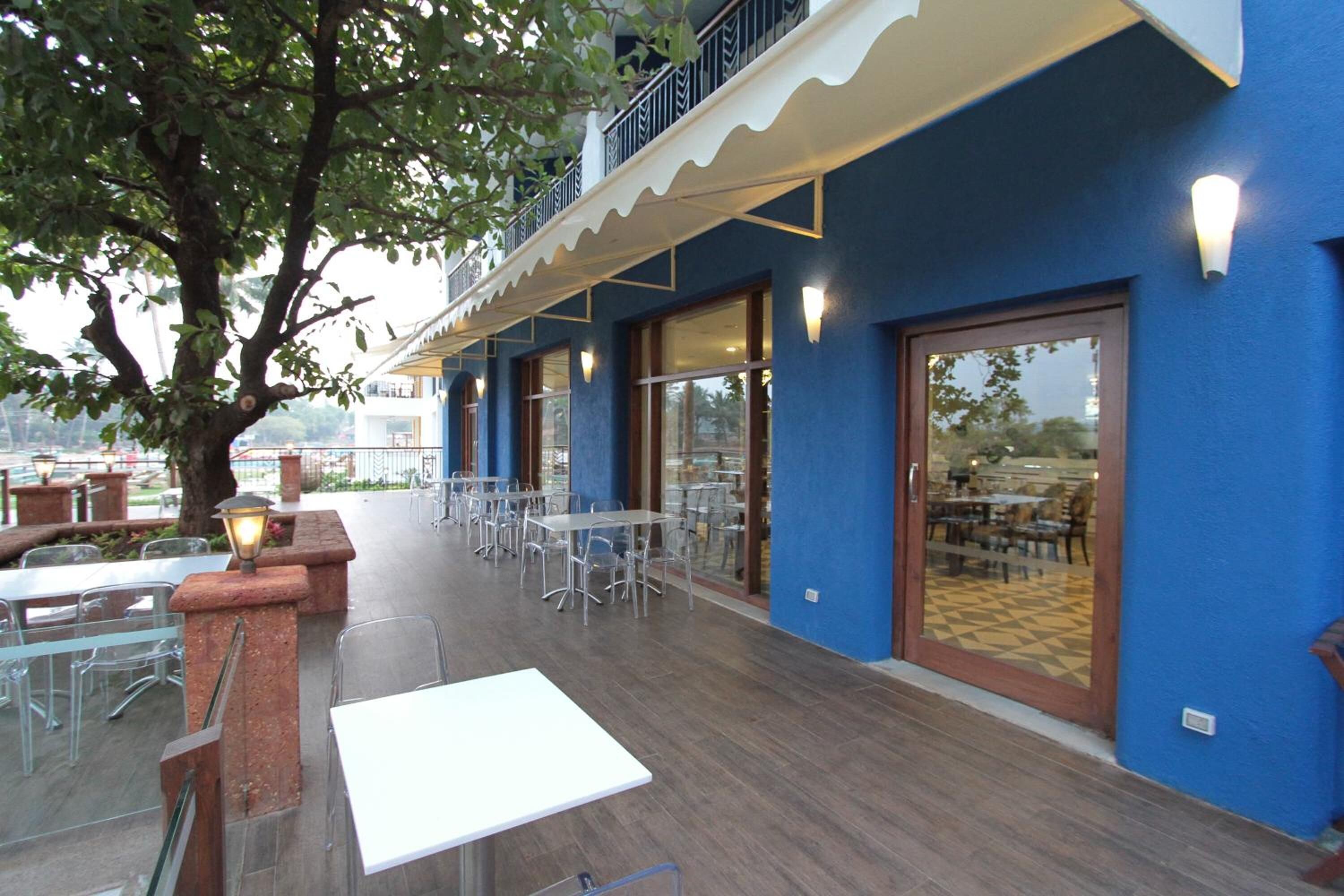 An outdoor dining area with tables and chairs, a tree, and a building with blue walls and a balcony.