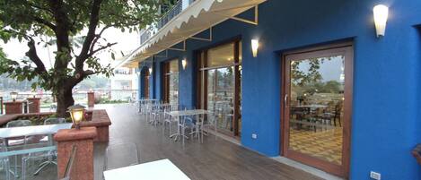 An outdoor dining area with tables and chairs, a tree, and a building with blue walls and a balcony.
