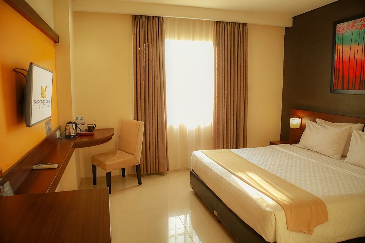 A hotel room with a bed, a desk with a chair, a TV, and a window with curtains.