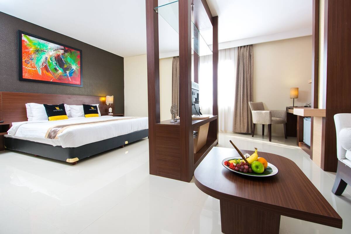 A hotel room with a bed, a desk with a fruit bowl, a chair, and a painting on the wall.
