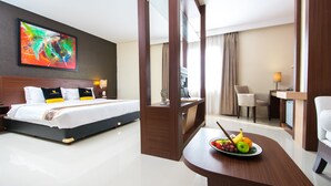 Una habitación de hotel con una cama, un escritorio con frutero, un televisor y un cuadro en la pared.