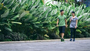 Dos personas caminando por un sendero pavimentado, con abundante follaje verde al fondo.