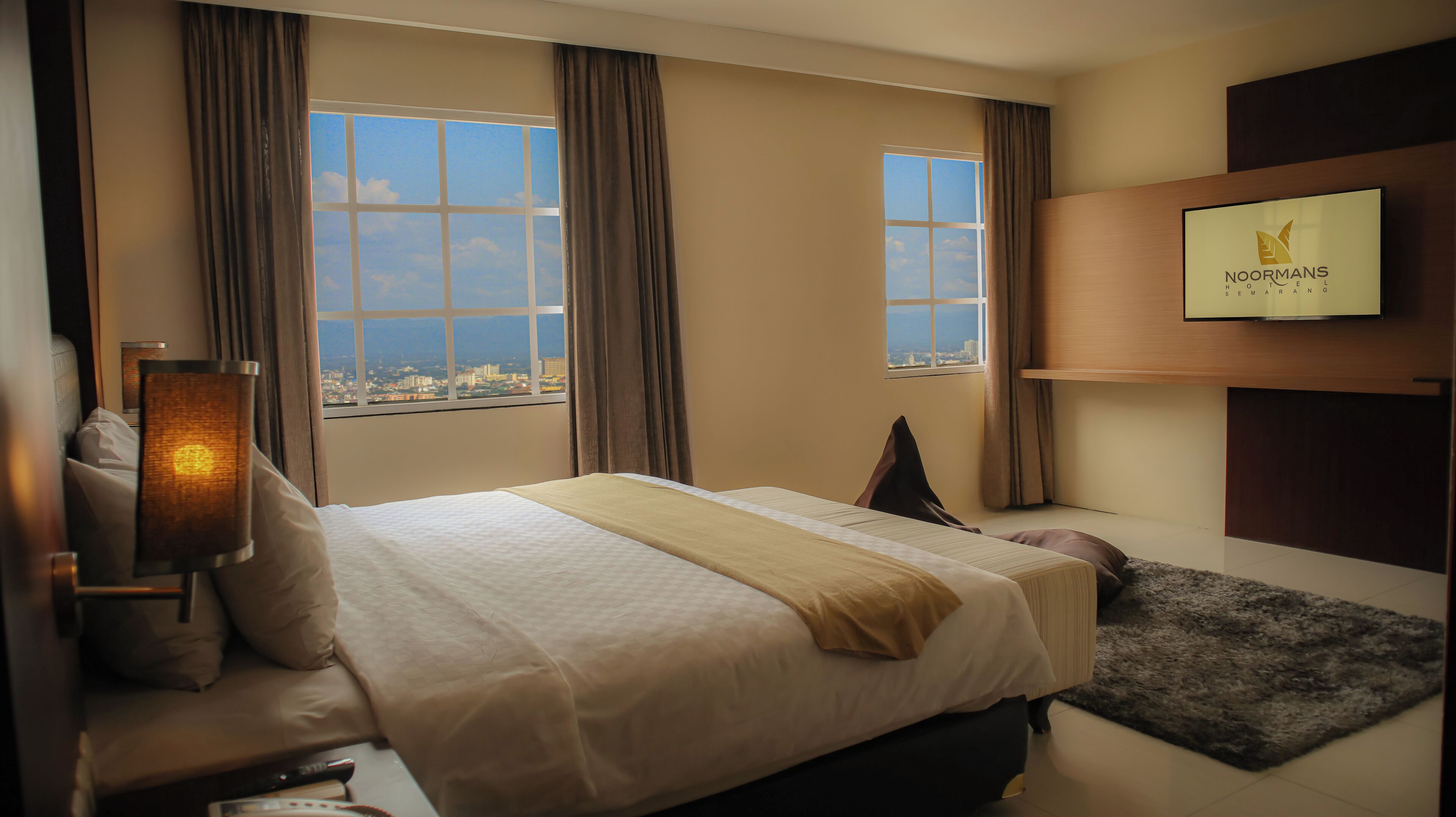 A hotel room with a large bed, a TV mounted on the wall, and a view of the city through the windows.