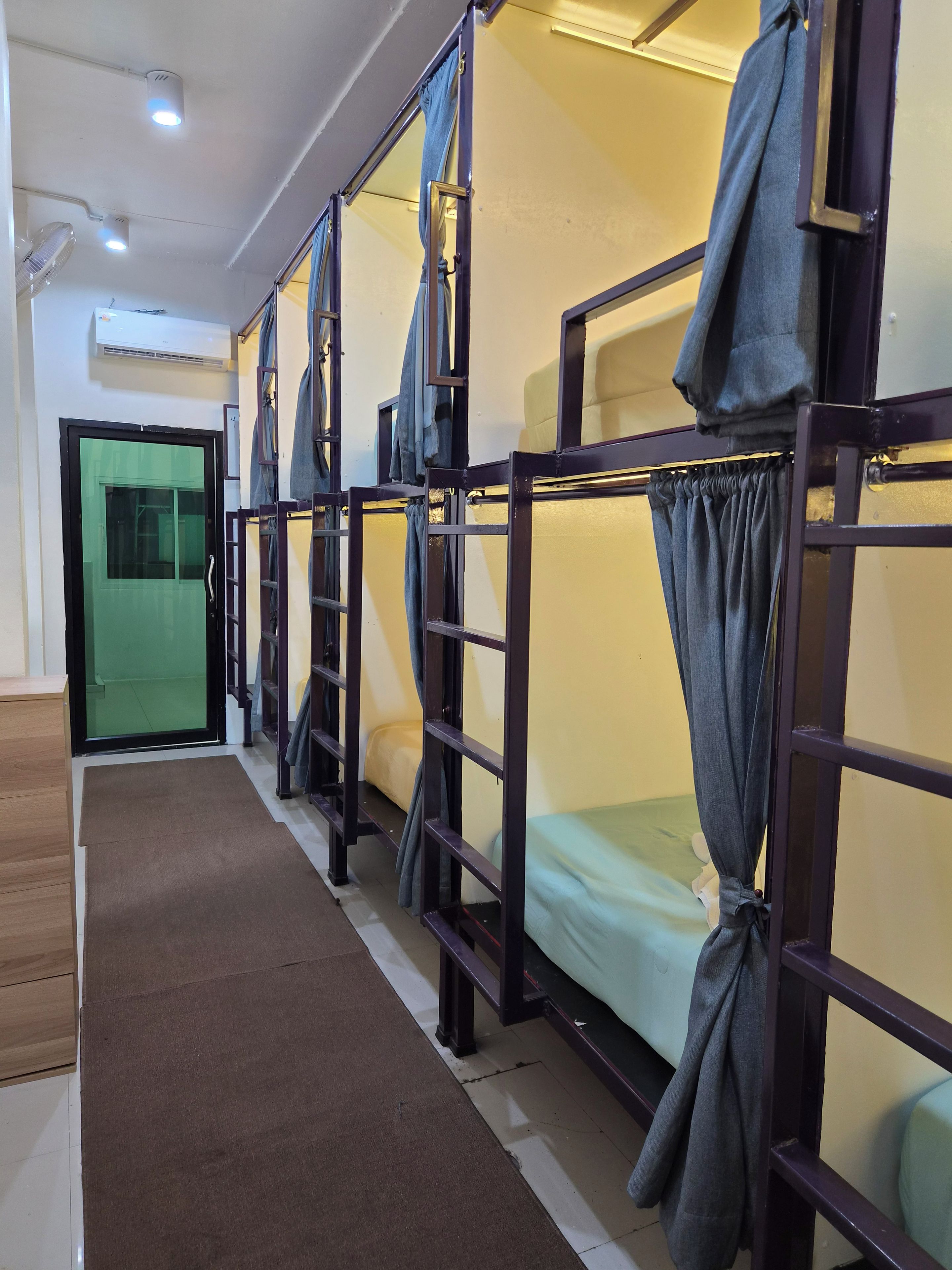 Une rangée de lits superposés avec des rideaux, une porte verte et un sol recouvert d’une moquette marron.