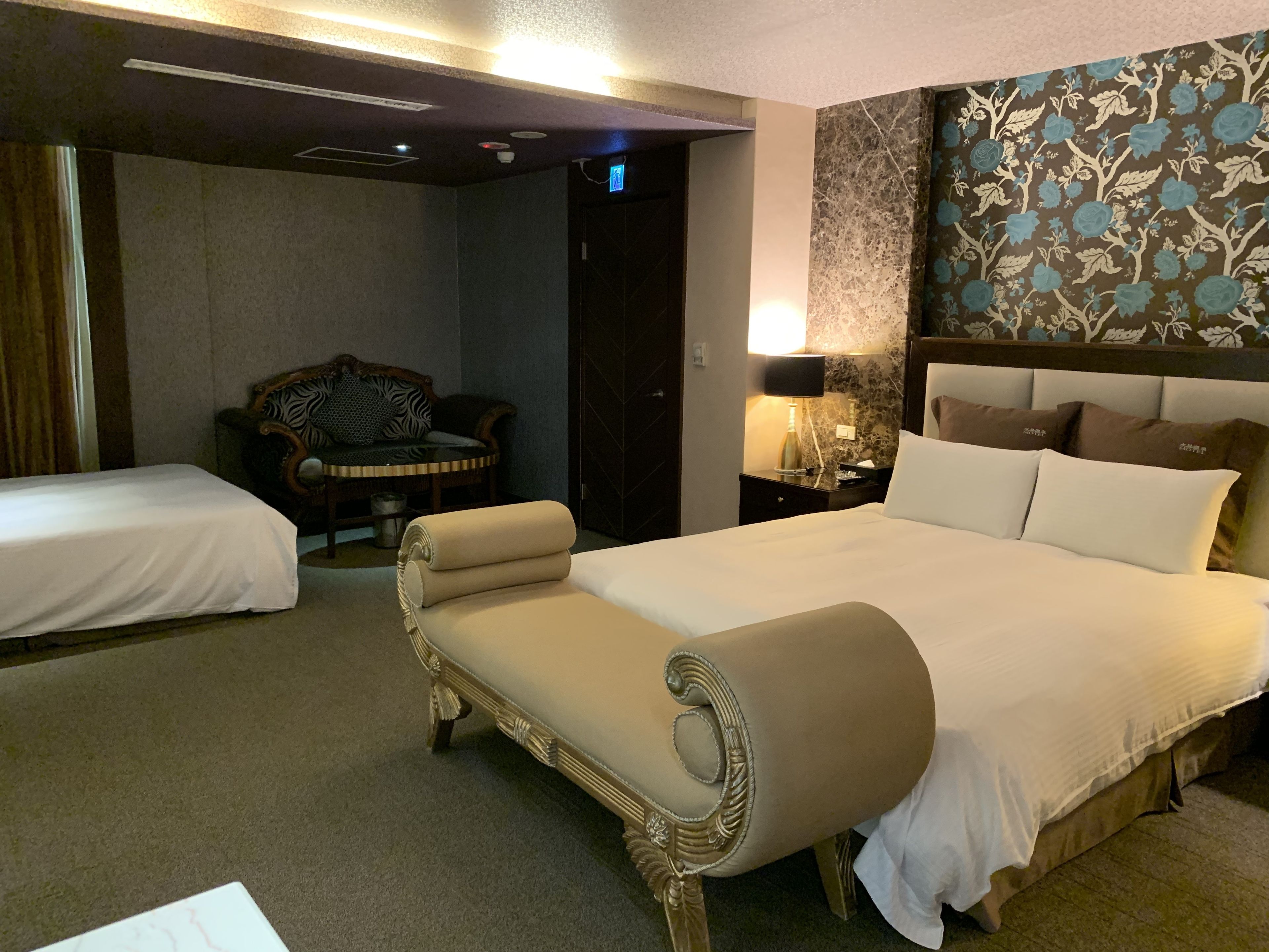 A hotel room with a large bed, a smaller bed, a chair, a nightstand with a lamp, and a wall with floral wallpaper.