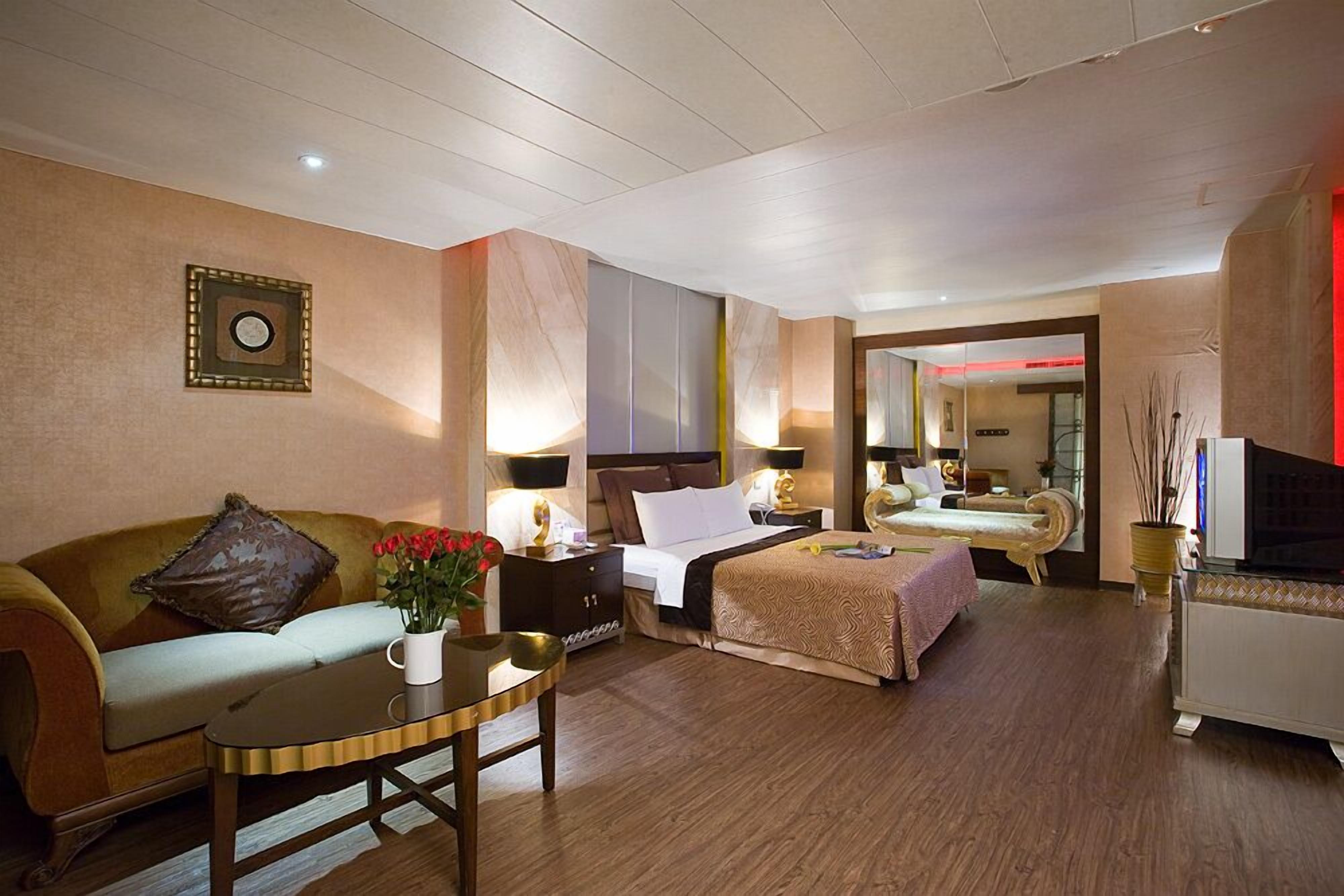 A hotel room with a sofa, a bed, a bedside table with a lamp, a TV, and a decorative vase with flowers.
