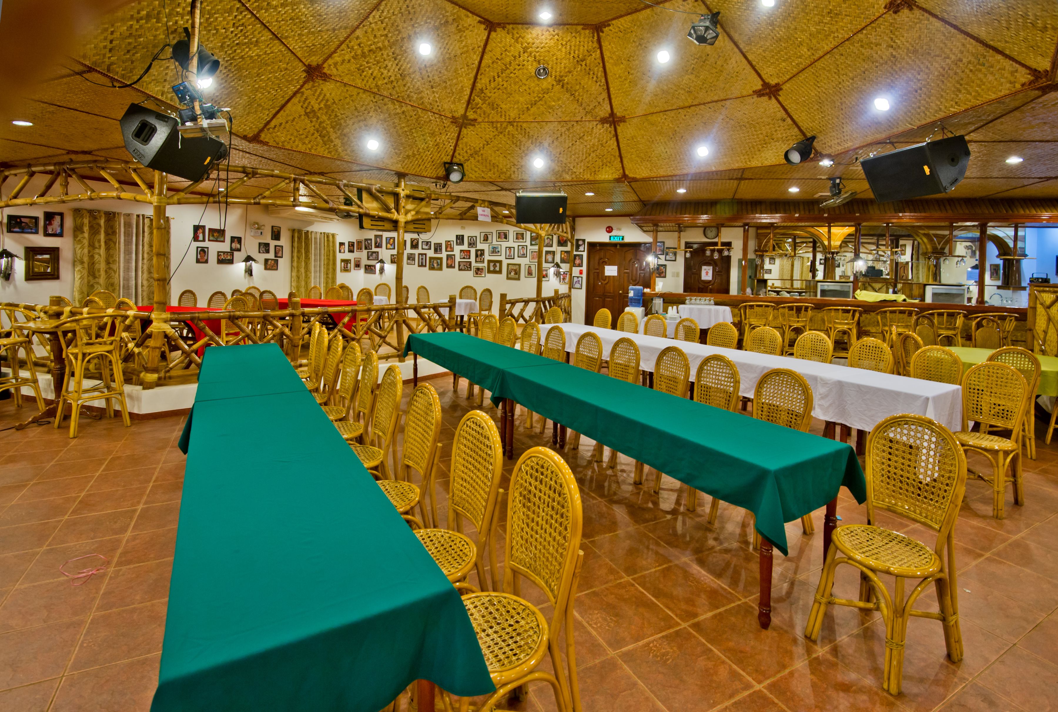 Un salón de banquetes con largas mesas cubiertas con manteles verdes, rodeado de sillas de mimbre.