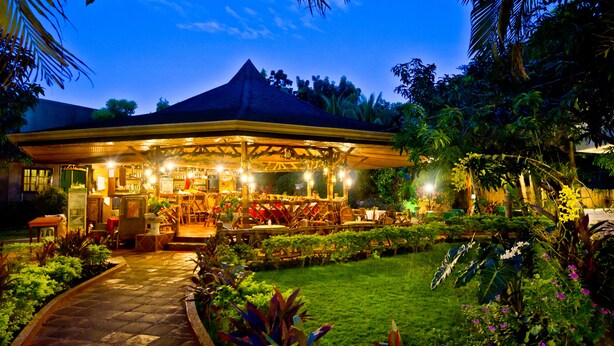 Un restaurant en plein air doté d’un espace repas couvert, entouré d’une végétation luxuriante et d’un jardin bien entretenu.