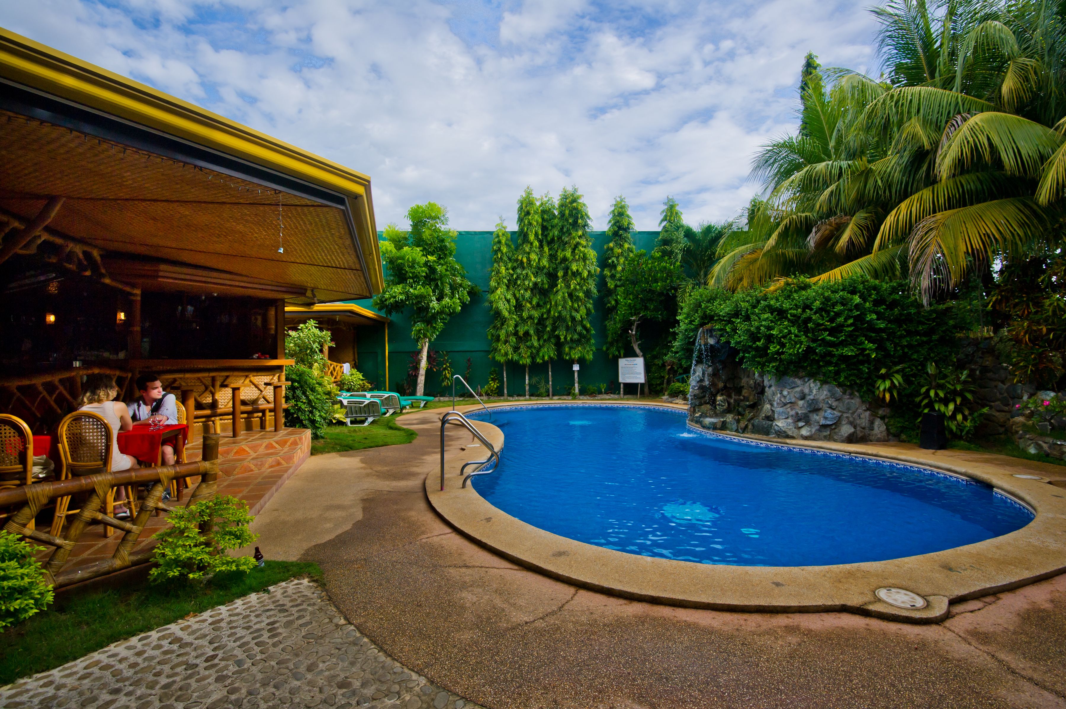 A pool area with a wooden deck, a restaurant with outdoor seating, and tropical vegetation.
