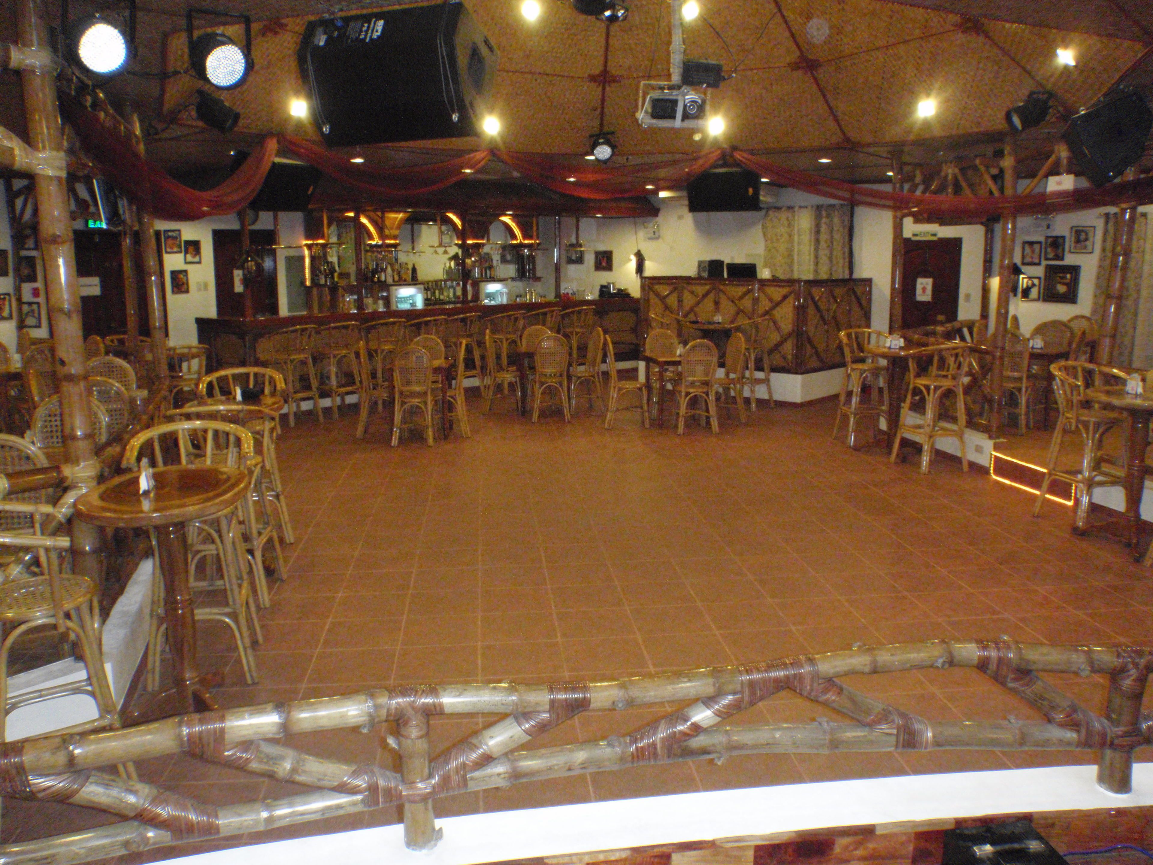 Un espacio interior amplio con numerosas sillas de mimbre dispuestas en filas, una zona de bar con taburetes y un escenario con un telón de fondo.