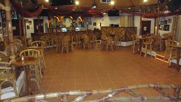 Un espacio interior amplio con numerosas sillas de mimbre dispuestas en filas, una zona de bar con taburetes y un escenario con un telón de fondo.