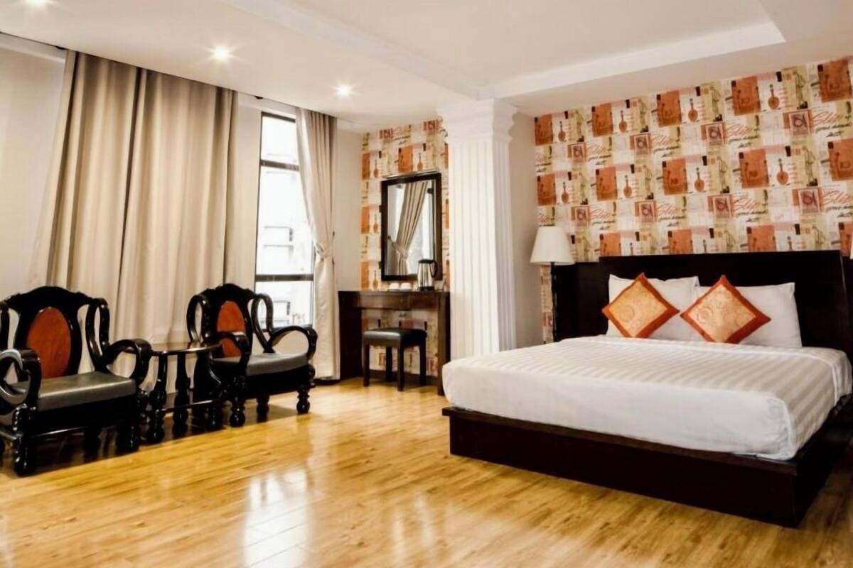 Une chambre d’hôtel avec un plancher en bois, un grand lit avec une literie blanche et deux coussins décoratifs, un bureau avec une chaise et une grande fenêtre avec des rideaux.