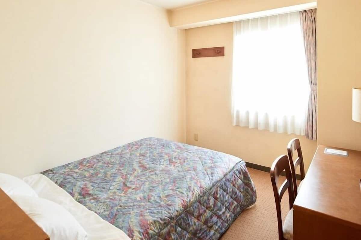Um quarto pequeno e simples, com uma cama, uma escrivaninha e uma janela com cortinas.