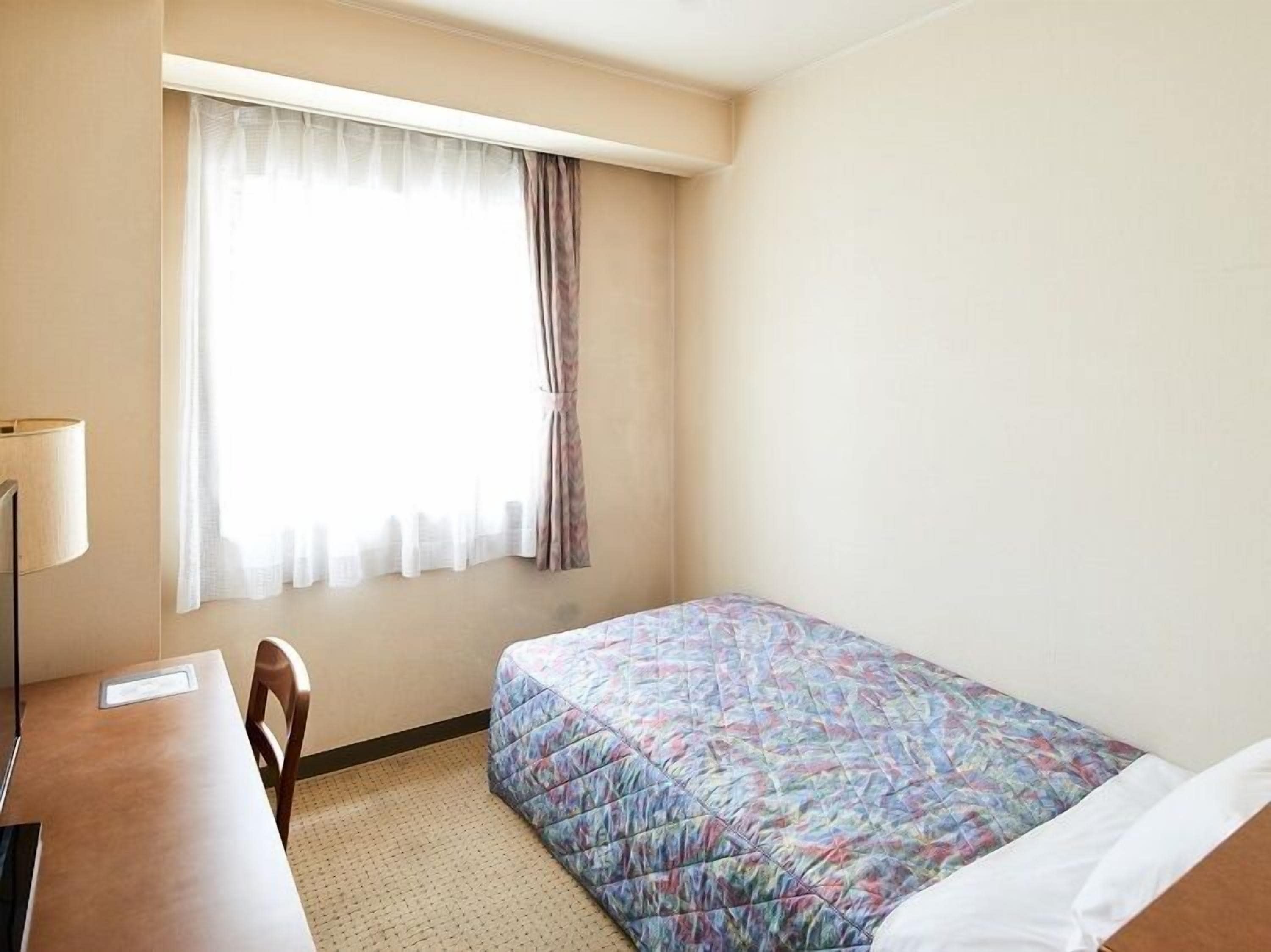 A hotel room with a bed, a desk, a chair, a window with curtains, and a lamp.