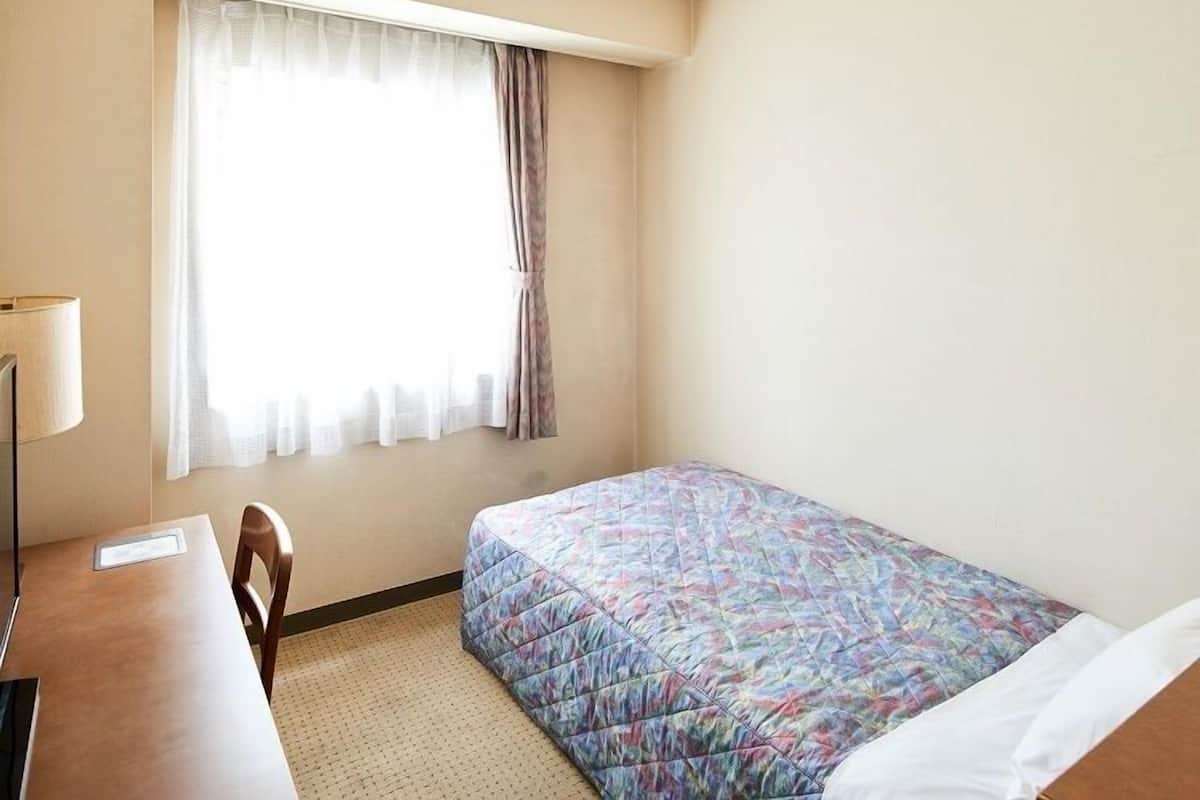 Quarto de hotel com uma cama, escrivaninha, cadeira, janela com cortinas e uma luminária.