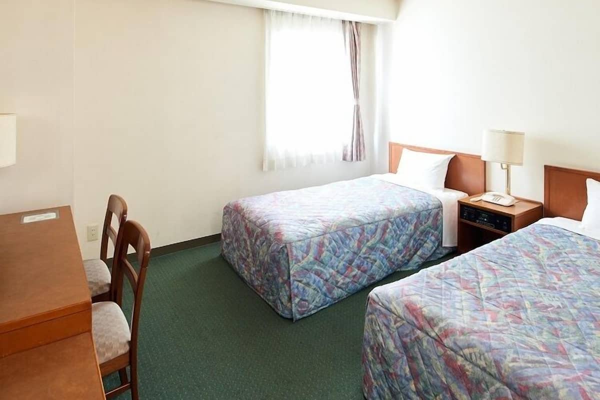 Quarto de hotel com duas camas, uma escrivaninha, uma cadeira, janela com cortinas e mesa de cabeceira com abajur.