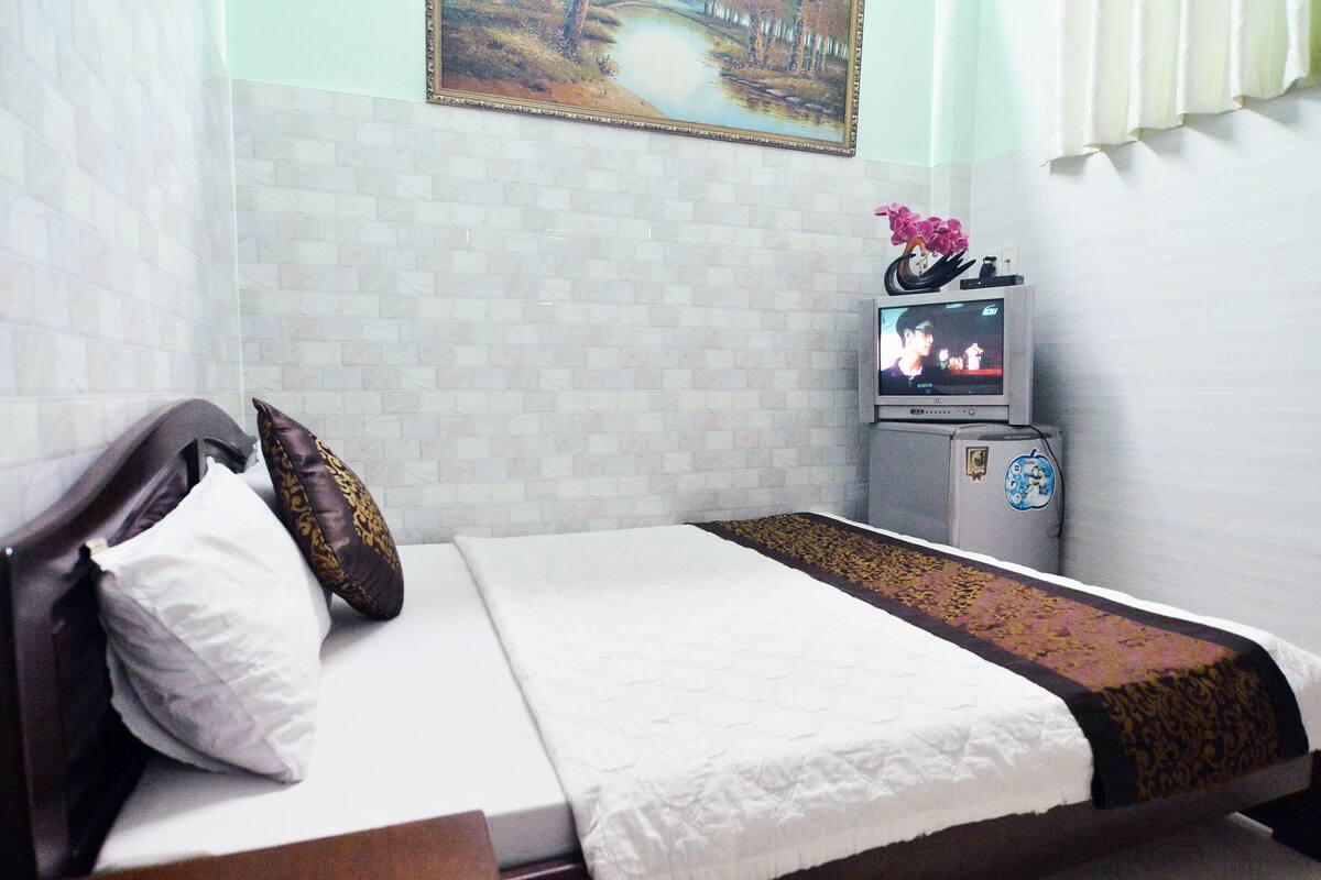 Una cama bien tendida con una colcha blanca y marrón, un almohadón blanco y uno marrón. Hay un televisor pequeño sobre un soporte con una decoración floral encima.