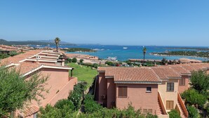 A coastal residential area with terracotta-roofed houses, a clear blue sky, and a view of the sea with boats.