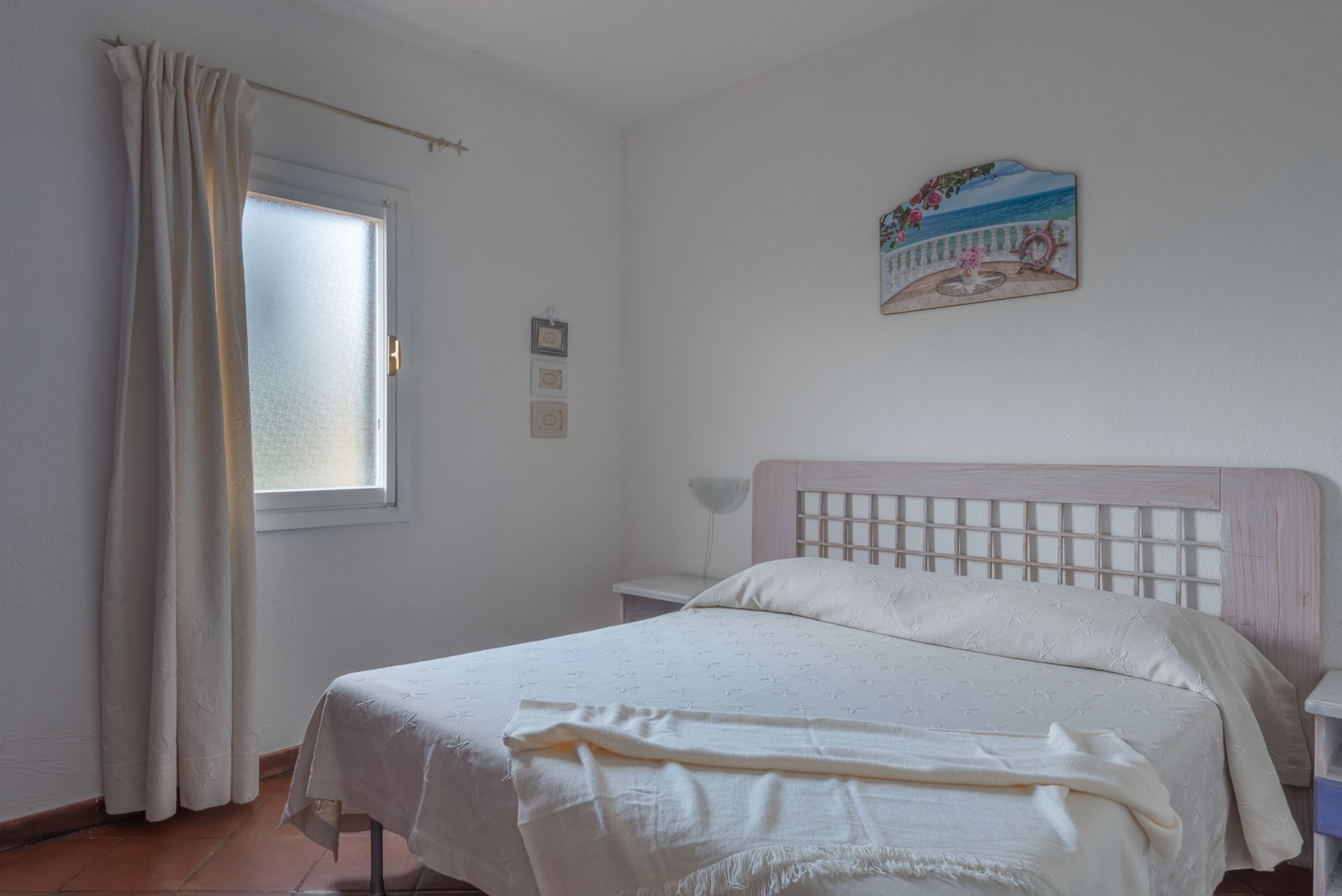 Ein Schlafzimmer mit Bett, einem Fenster mit Vorhängen, einem Nachttisch und einem an der Wand hängenden Bild.
