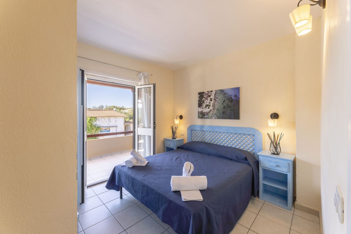 Une chambre à coucher avec un lit, une table de chevet, une lampe et une vue sur l’extérieur.