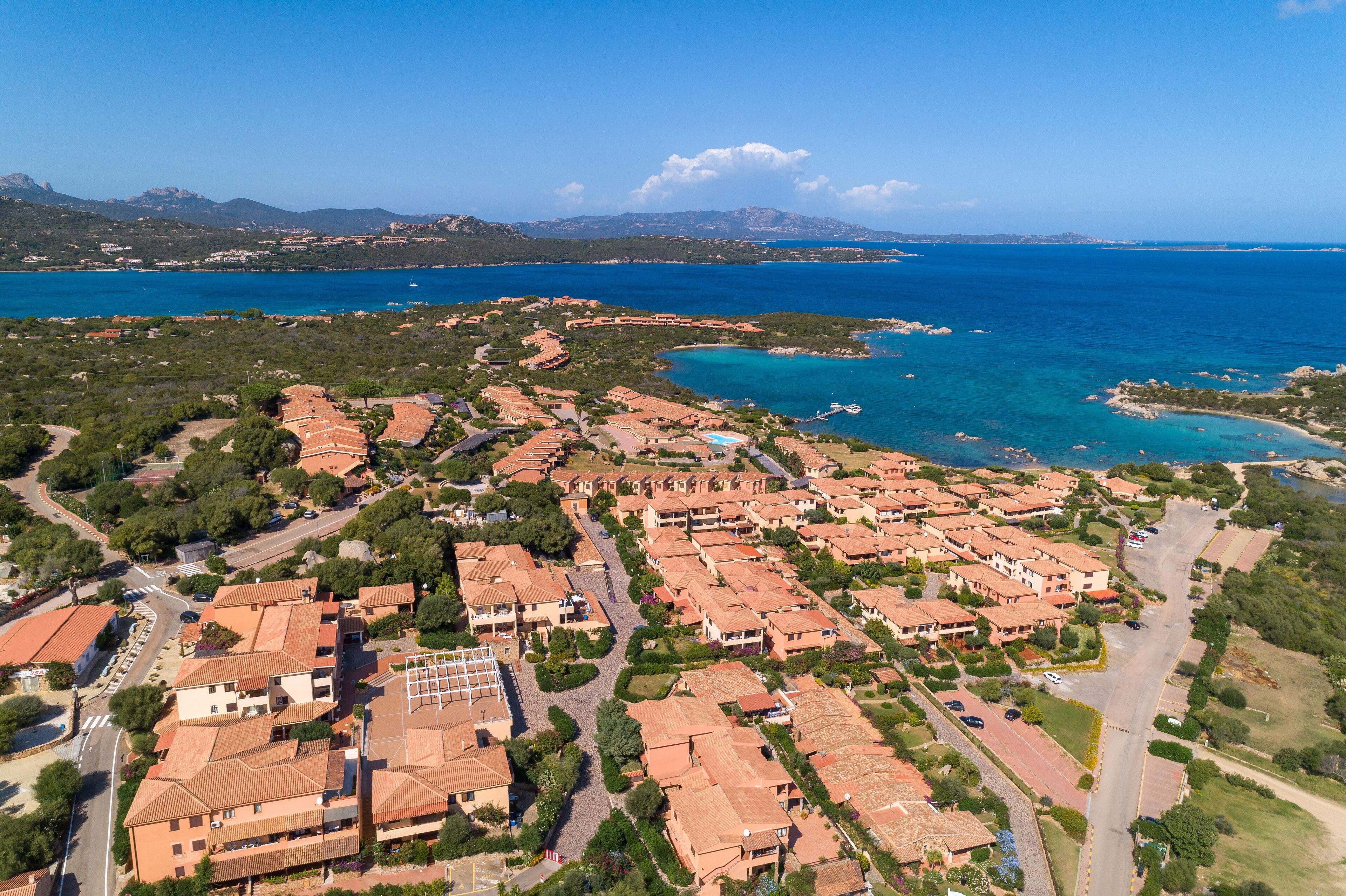 An aerial view of a coastal residential area with terraced houses, roads, and a clear view of the sea and distant mountains.