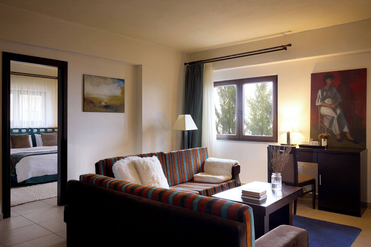 A hotel room with a striped sofa, a small table with a lamp, a painting, and a view of the outdoors through a window.