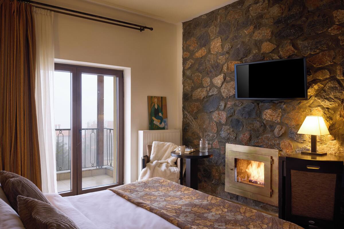 A hotel room with a bed, a fireplace, a television mounted on a stone wall, and a balcony with a view.