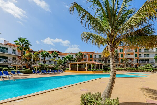 A swimming pool with lounge chairs and a palm tree, surrounded by multi-story buildings with balconies. - Agua Hotels Sal Vila Verde Resort (Sal)