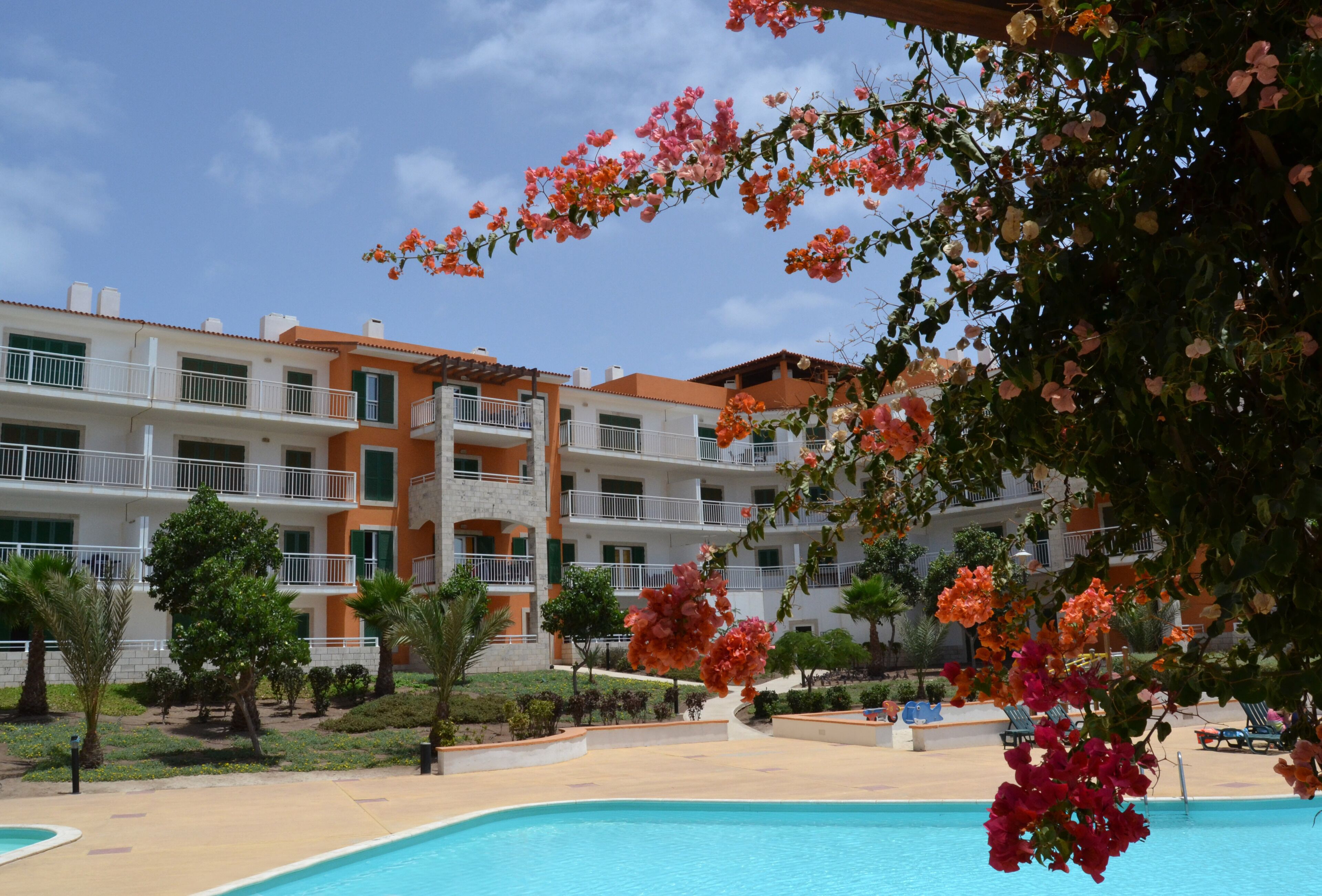 A hotel with a pool, surrounded by greenery and a clear blue sky.