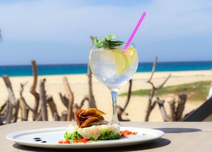 A glass of iced drink with a lime slice and mint garnish on a white plate, accompanied by a piece of fried food, on a table with a beach view in the background. - Agua Hotels Sal Vila Verde Resort (Sal)